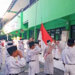 Tagar.co – Barisan siswa berseragam merah putih, bertopi warna senada dan berdasi merah terlihat rapi. Gerakan mereka selaras dengan panduan pembina upacara yang memimpin jalannya upacara di halaman SD Negeri Bulak Rukem 1 Surabaya, Senin (29/7/2024). Mereka sudah masuk di area lapangan sejak pukul 6.15 WIB. Upacara bendera tiap Senin itu dimulai tepat ketika jarum jam menunjukkan pukul 6.30 WIB. Tepat ketika hendak mulai, tiba-tiba dua anak laki-laki berlarian pontang panting menuju barisan. Sambil terengah-engah, mereka membetulkan letak dasi dan topinya. Ialah Azel Fedora Isatitaah (10) dan Barnes Fasle Maula (10). Tak mereka hiraukan peserta upacara yang memandang dengan penuh tanya. Begitupun dengan pandangan para guru yang turut berdiri di barisan depan siswa, berdampingan dengan para petugas upacara berseragam putih di barisan depan. Pemeriksaan kelengkapan atribut sekolah menjadi awal kesiapan barisan yang dipimpin oleh pemimpin barisan. Harapannya, anggota memeriksa kelengkapan seragam sekolahnya. Mulai dari topi, dasi, baju putih dengan badge kelas di lengan kanan, nama lengkap di dada kanan dan logo Tut Wuri Handayani di saku kiri. Selain itu, memakai ikat pinggang berlogo SD. Juga bawahan rok untuk siswa perempuan dan celana untuk siswa laki-laki. Berakhir pada kaos kaki putih dan sepatu hitam. Para siswa memeriksa semua itu sesuai instruksi Chairul Anwar, Wali Kelas VI E. Dia yang pagi itu mendapat amanah mengatur kesiapan peserta sebelum upacara resmi dimulai. "Hari ini yang atributnya belum lengkap masih kita beri waktu untuk dilengkapi hingga Senin depan. Bagi yang belum punya, bisa beli di koperasi sekolah agar seragam kalian rapi," pesannya di tengah depan lapangan. Sejenak suasana hening ketika Achmad Maulana Rahmatullah (11), bertugas sebagai pemimpin upacara, menyiapkan barisan. "Seluruhnya, siap grak!" tegasnya lantang. Satu per satu petugas upacara menjalankan tugasnya. Hingga tiba giliran Paskibraka mengibarkan sang Merah Putih dengan kompak. Mereka menjalankan tugasnya sampai bendera Indonesia berkibar di angkasa. Santri Surabaya Juara FASI XII Diterima Sekda Disiplin dari Rumah Upacara hari Senin yang sudah menjadi rutinitas sekolah itu punya peserta baru. Mereka yang sebelumnya kelas IV masuk siang, kini naik kelas V dan masuk pagi. Jadi ada perubahan pembiasaan yang membuat siswa perlu menyiapkan diri. Pagi itu, yang menjadi pembina upacara ialah Masbuchin, guru mata pelajaran Pendidikan Agama Islam dan Budi pekerti. Kesempatan menyampaikan amanat upacara ia manfaatkan untuk menekankan pesan moral terkait kedisiplinan. "Selamat untuk siswa kelas IV yang naik kelas V dan yang kelas V naik kelas VI. Khususnya kelas V yang semula di kelas IV masuk siang, sekarang di kelas V masuk pagi," sapanya di awal. Kepada siswa kelas V, Masbuchin mengharuskan mereka mulai menata pola tidur dan disiplin waktu. Harapannya, mereka bisa datang tepat waktu ke sekolah. Lebih lanjut, ia mengatakan, "Mulai hari ini, setiap Senin akan ada upacara. Maka sejak pukul 6.30 seluruh siswa sudah harus di lapangan berbaris rapi untuk melatih kedisiplinan dan kemandirian kalian." Latihan kedisiplinan ini menurutnya dimulai dari menyiapkan diri ketika di rumah. "Berawal dari istirahat yang cukup, menyiapkan jadwal pelajaran, dan seragam sekolah malam hari di rumah," urainya. Dengan disiplin sejak di rumah, ia meyakini, esok paginya anak-anak lebih siap ke sekolah. (#) Jurnalis Tri Eko Sulistiowati Penyunting Sayyidah Nuriyah